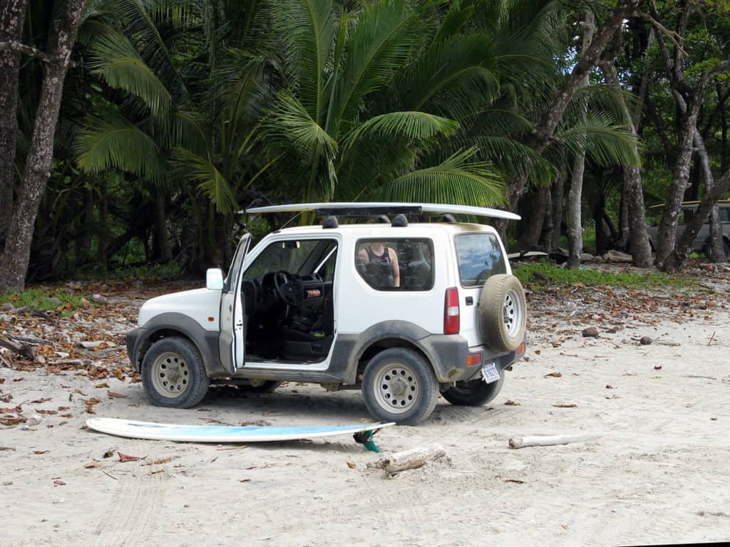 A white suv parked on the sand in Costa Rica with a surfboard.