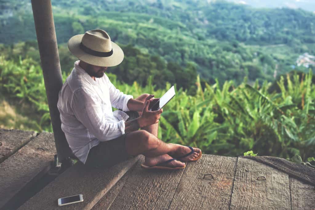 A man in a hat sits on a ledge in Costa Rica while using a tablet.